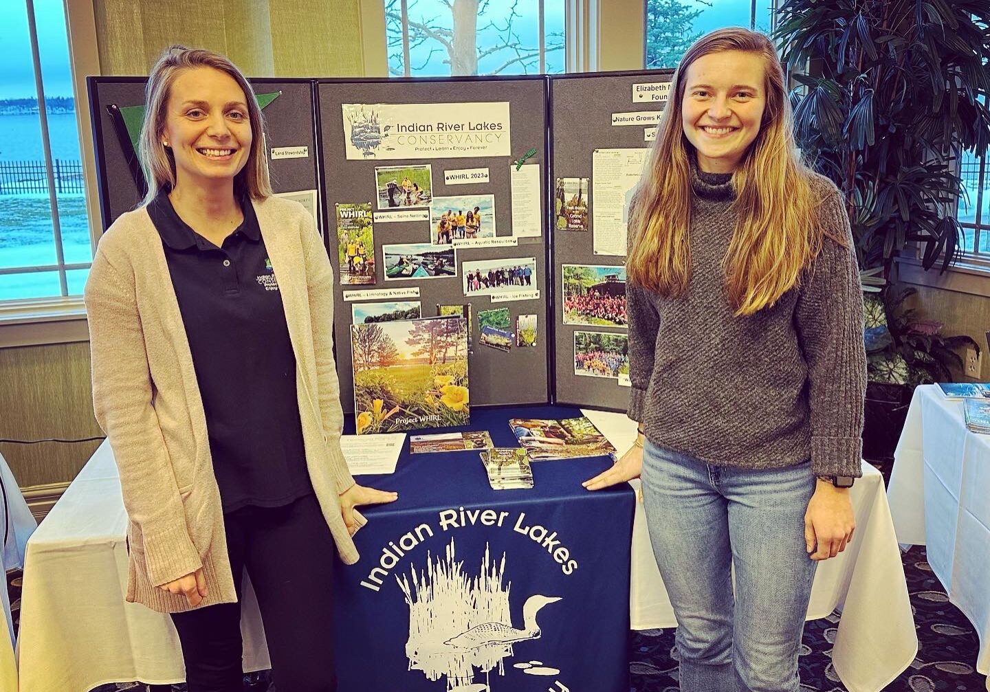 Two Women stand in front of an IRLC display table